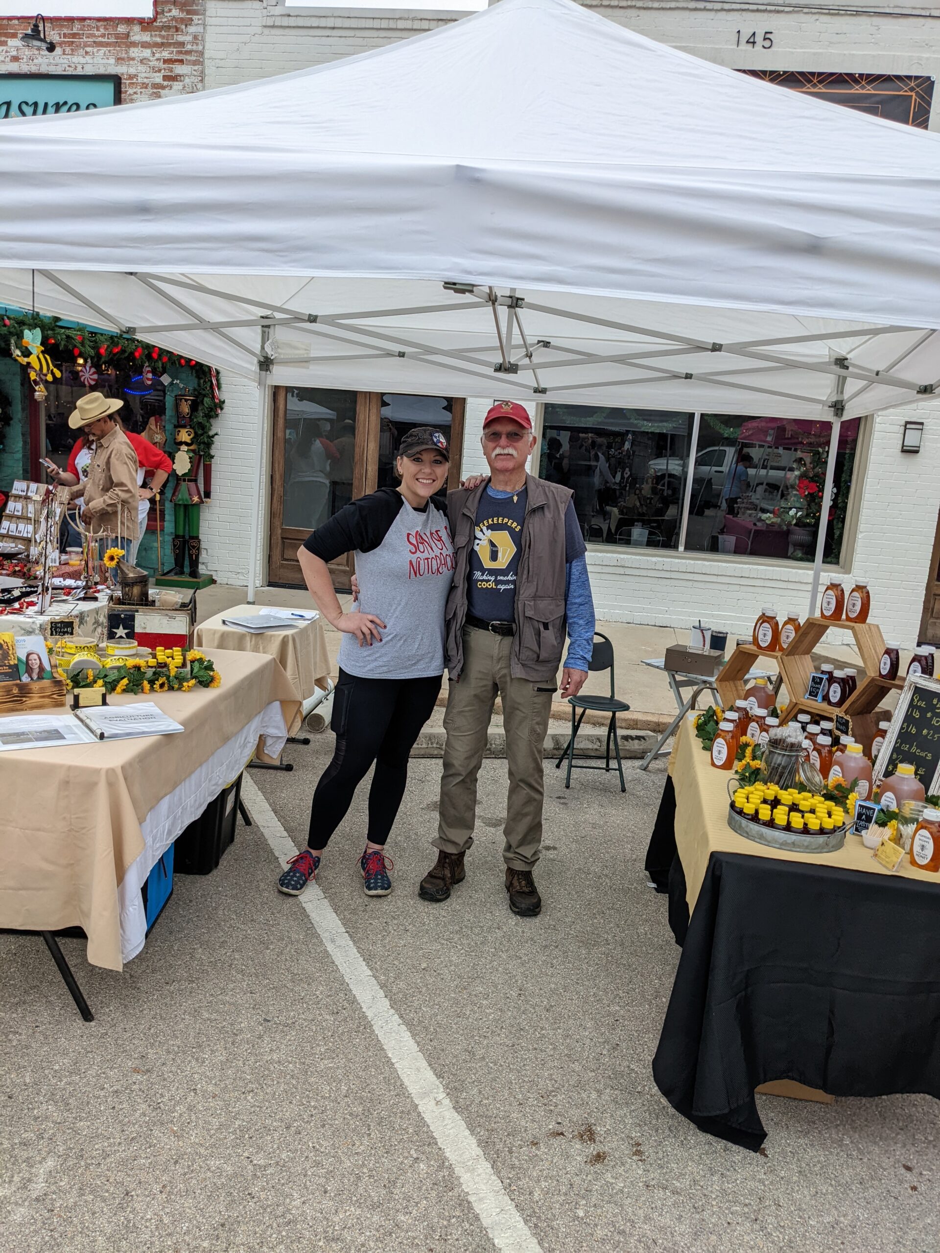 A smiling man and woman stand under a white canopy at a market. Tables around them display honey products and decorations. Honeycomb graphics frame the image.