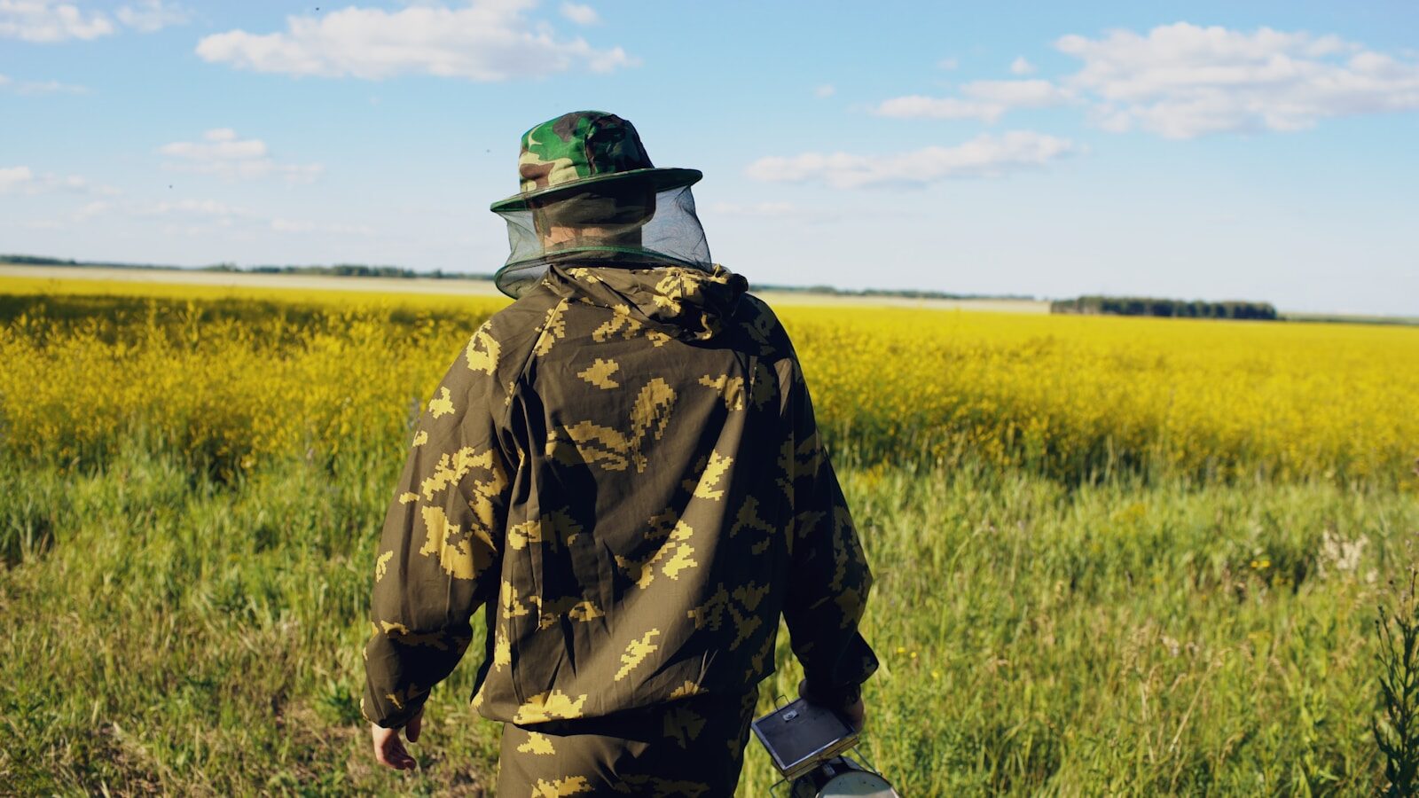 Beekeeper in protective suit walks through a yellow flower field.